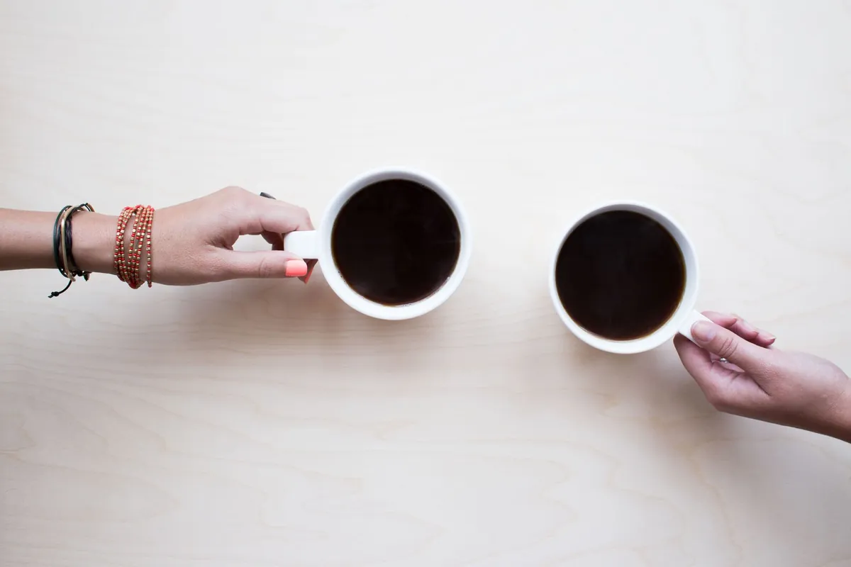 Two hands reaching for two identical cups of coffee on a white surface