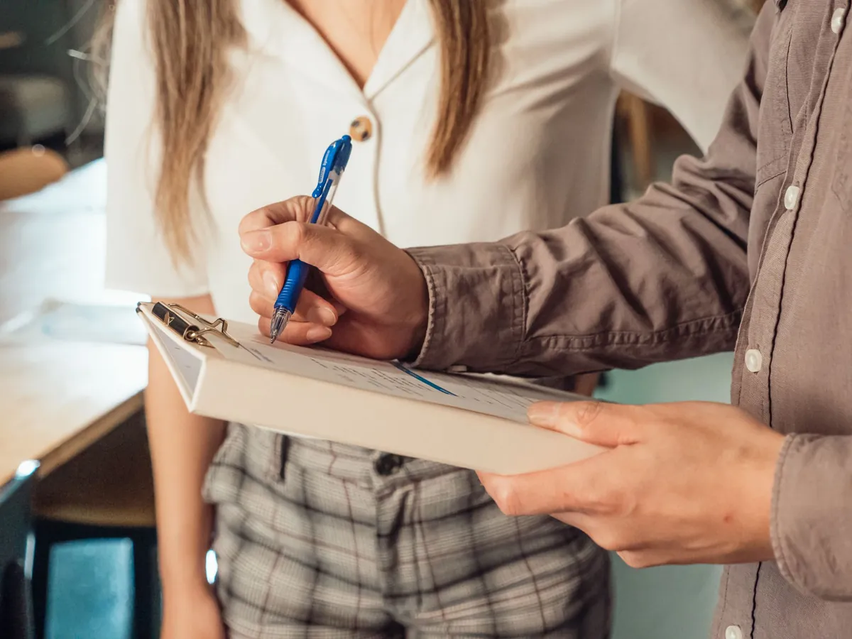 One person writing on a clipboard while another holds it, in an office