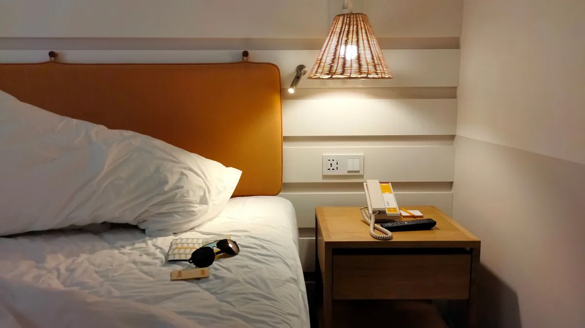 A cream corded telephone handset resting on a wooden bedside table next to a small notepad and pencil, beside a bed with a white duvet under a lamp.