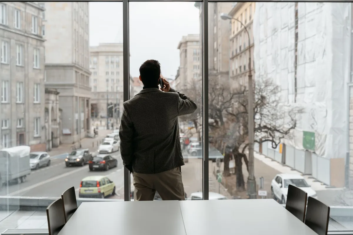 A man in a tweed jacket stands with his back to the camera at a floor-to-ceiling window, phone held to his ear, looking down on a city street with traffic.