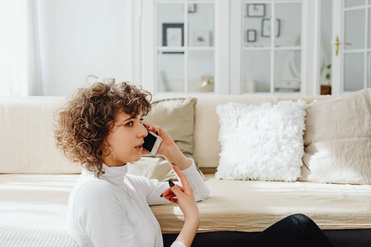 A woman on a phone call sitting on the floor against a white sofa