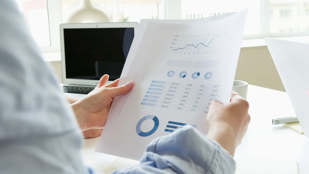 A person holding a printed chart and reading it beside an open laptop