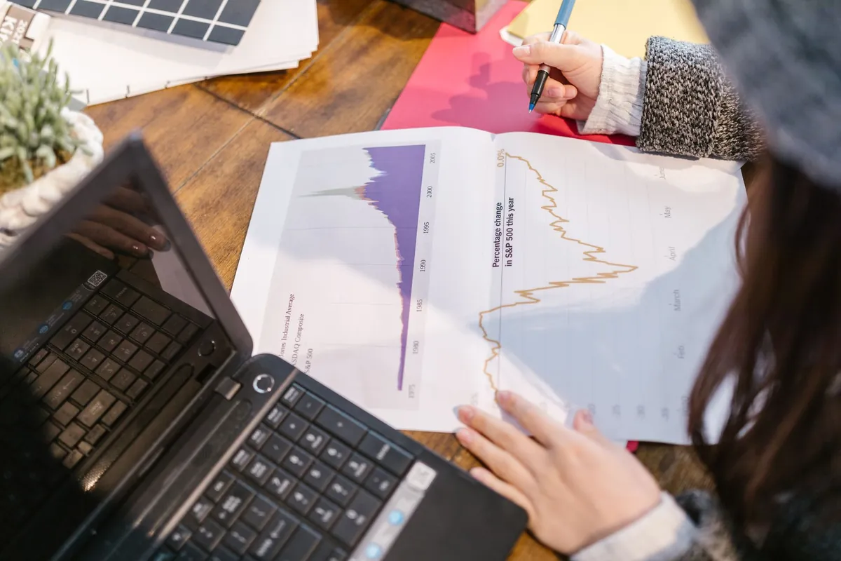 A person reading a printed graph and taking notes beside a laptop