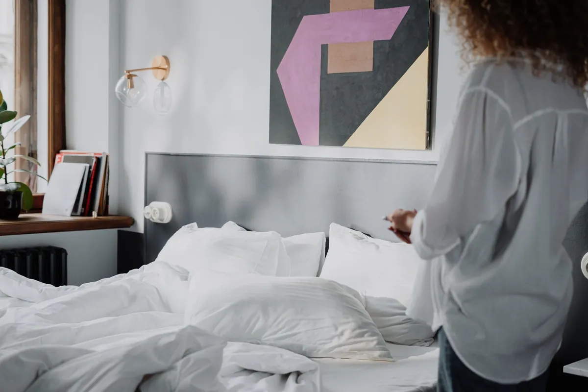 Person in a light shirt standing beside an unmade white bed in a bright bedroom