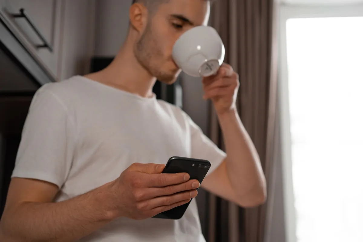 Young man in a white t-shirt drinking from a mug while looking at his phone