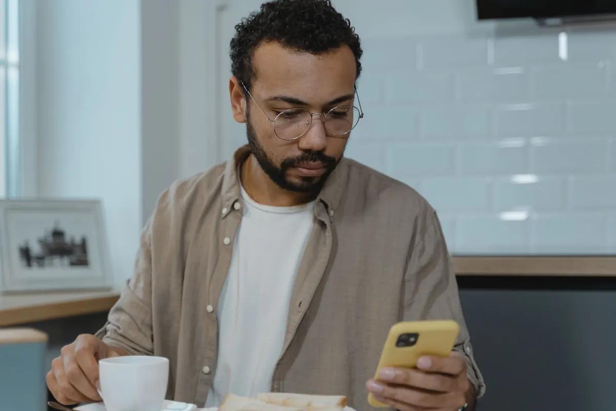 A man wearing eyeglasses checks his smartphone while eating breakfast indoors.