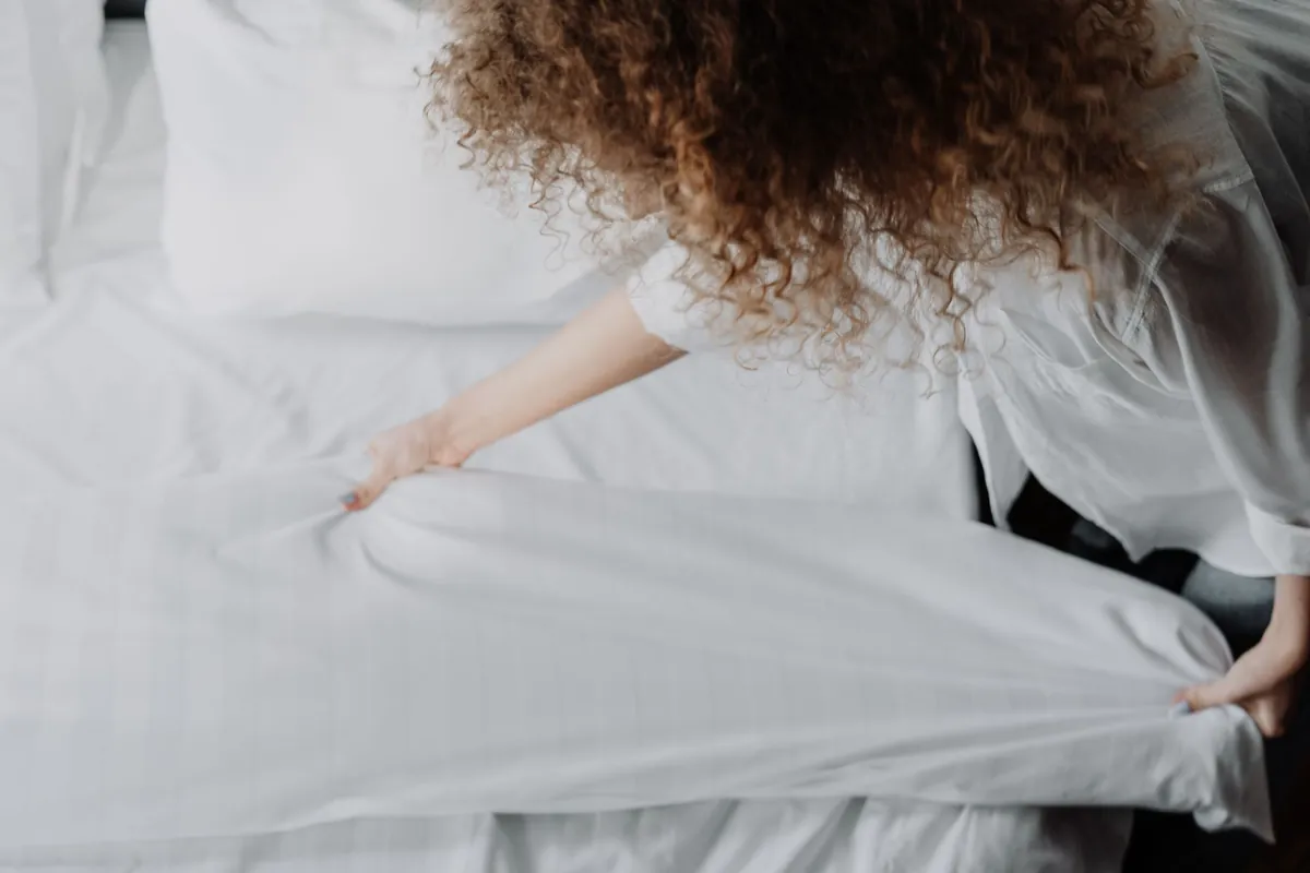 A person smoothing white bed linens on a bed in a calm, tidy bedroom