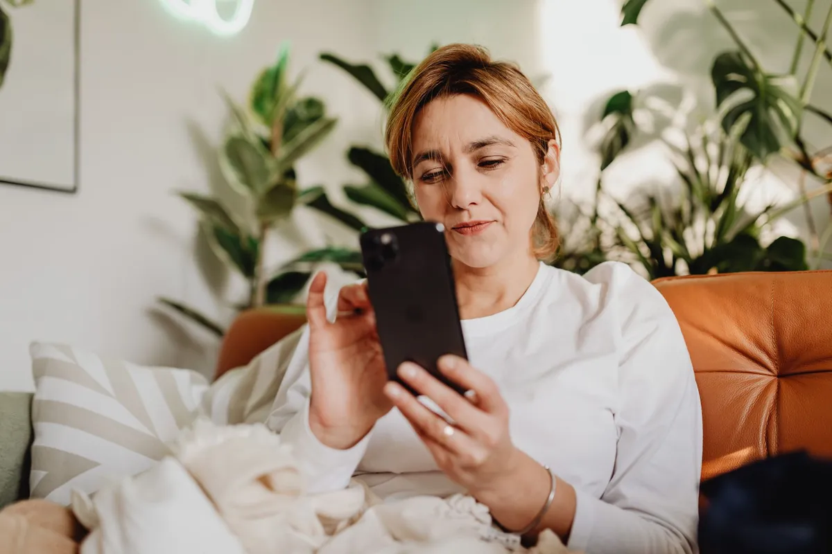 A woman sitting on a cozy sofa at home, using her smartphone, with houseplants around her