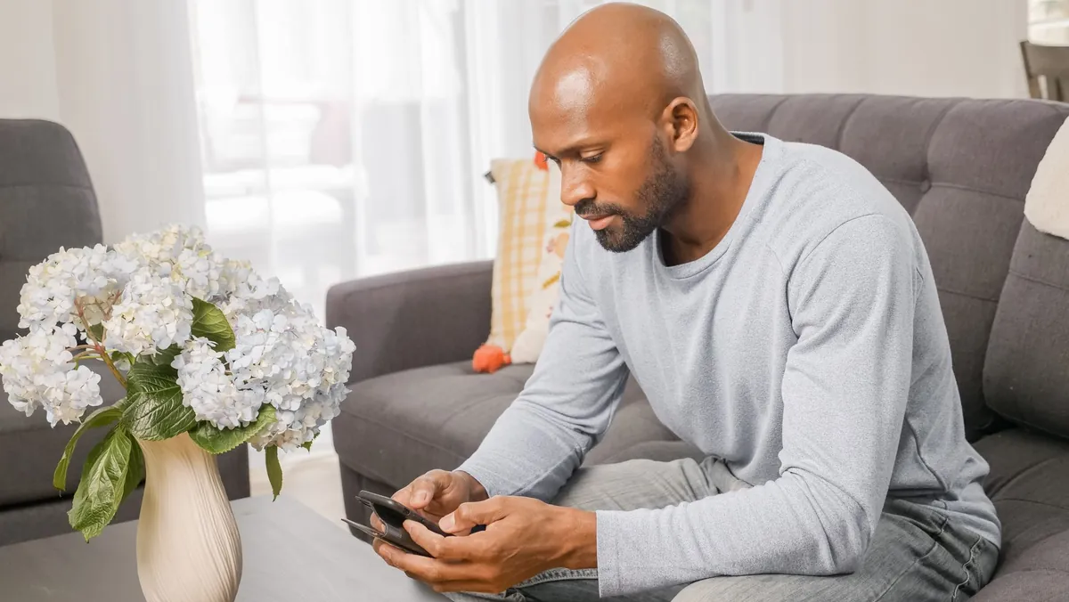Man sitting on a couch at home looking at his smartphone beside a vase of white flowers