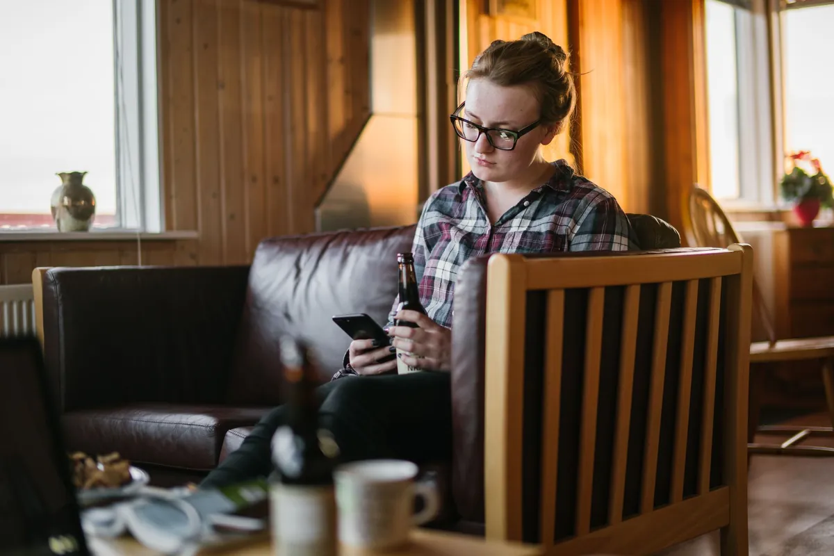 Woman sitting on a sofa indoors, holding a drink and looking at her smartphone