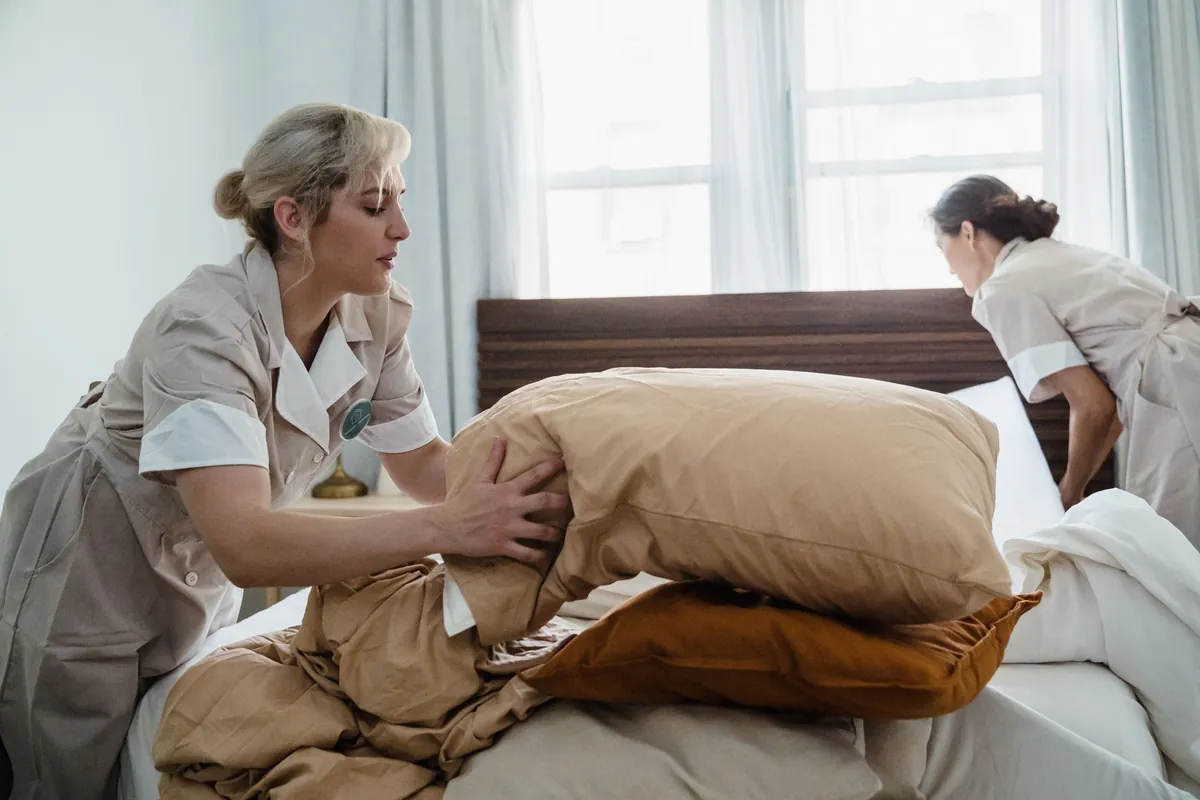 Two housekeepers in beige uniforms making a bed and arranging pillows by a bright window