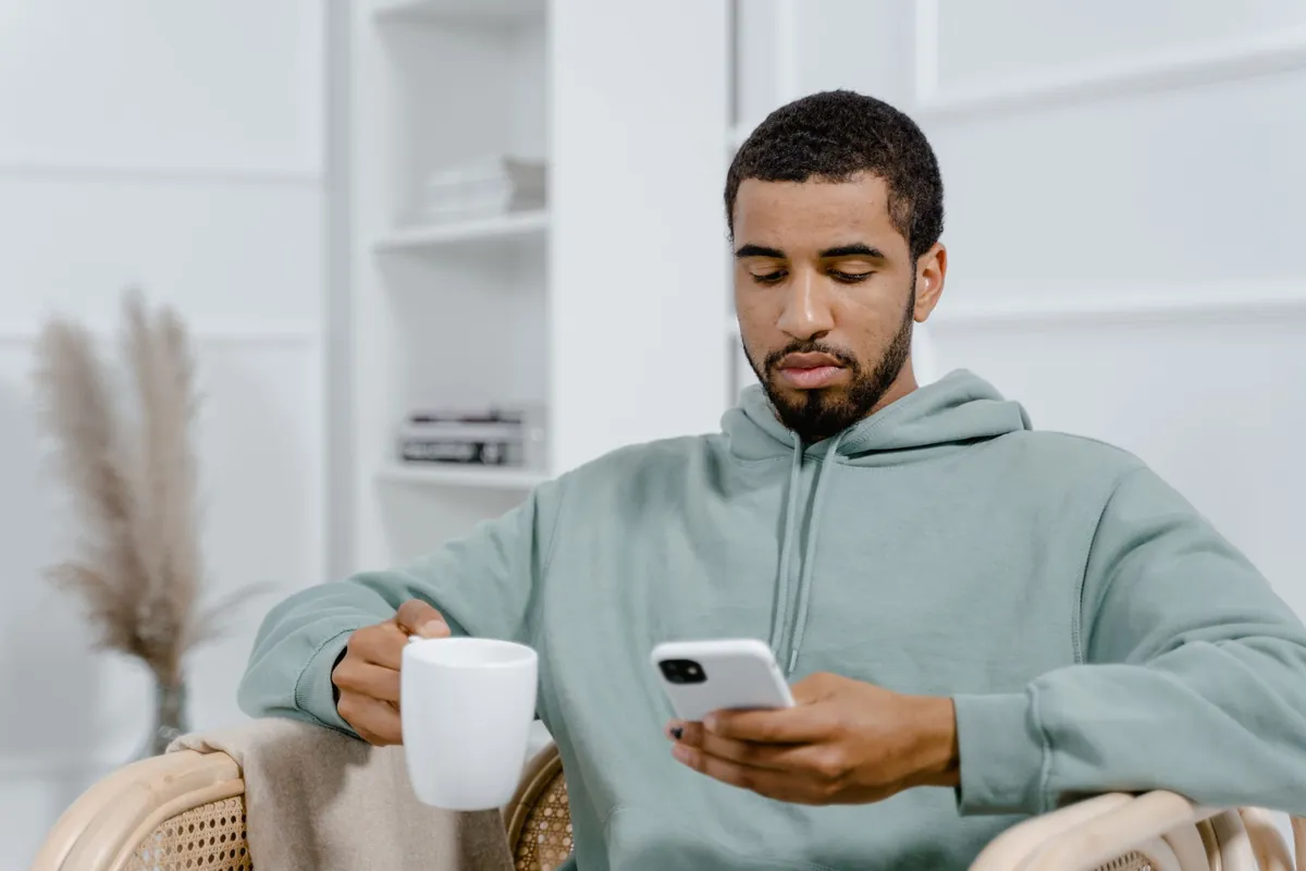 A man sitting in a cozy living space using a smartphone while holding a coffee mug.
