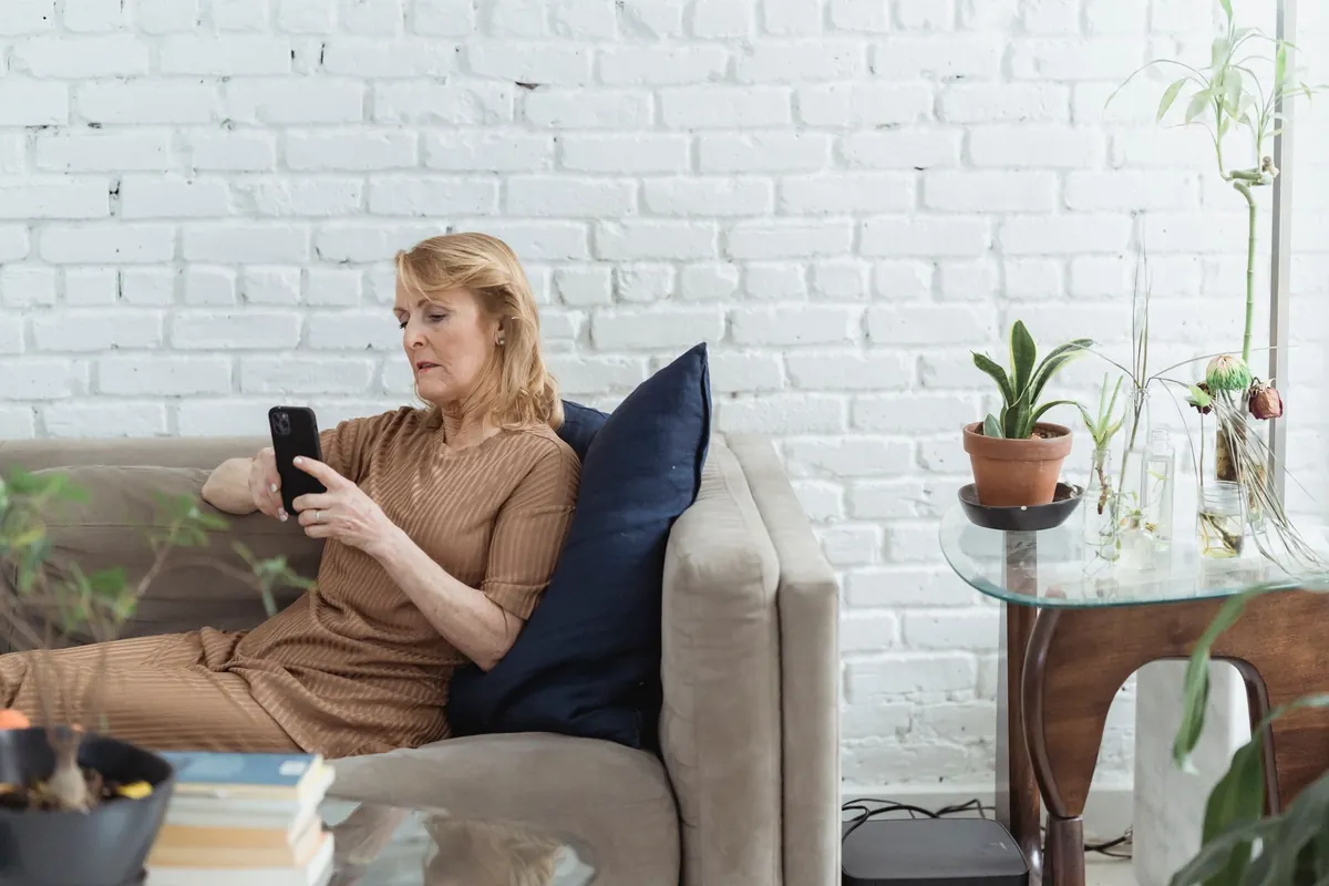 Senior woman browsing the internet on her cellphone while resting on a couch near brick wall and potted plants.