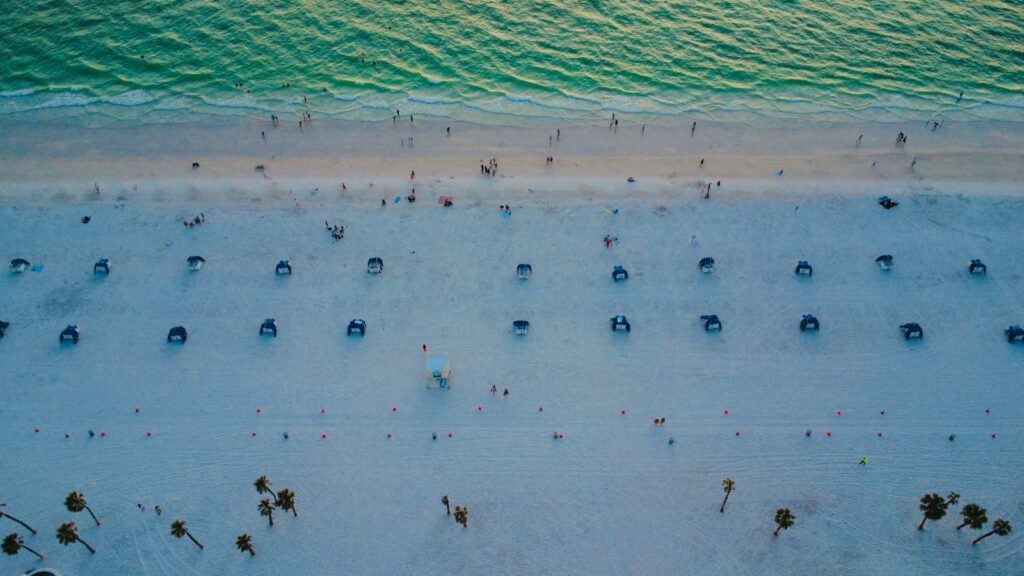 Aerial view of a white sand beach in Florida with rows of beach cabanas, palm trees, and green water