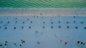 Aerial view of a white sand beach in Florida with rows of beach cabanas, palm trees, and green water