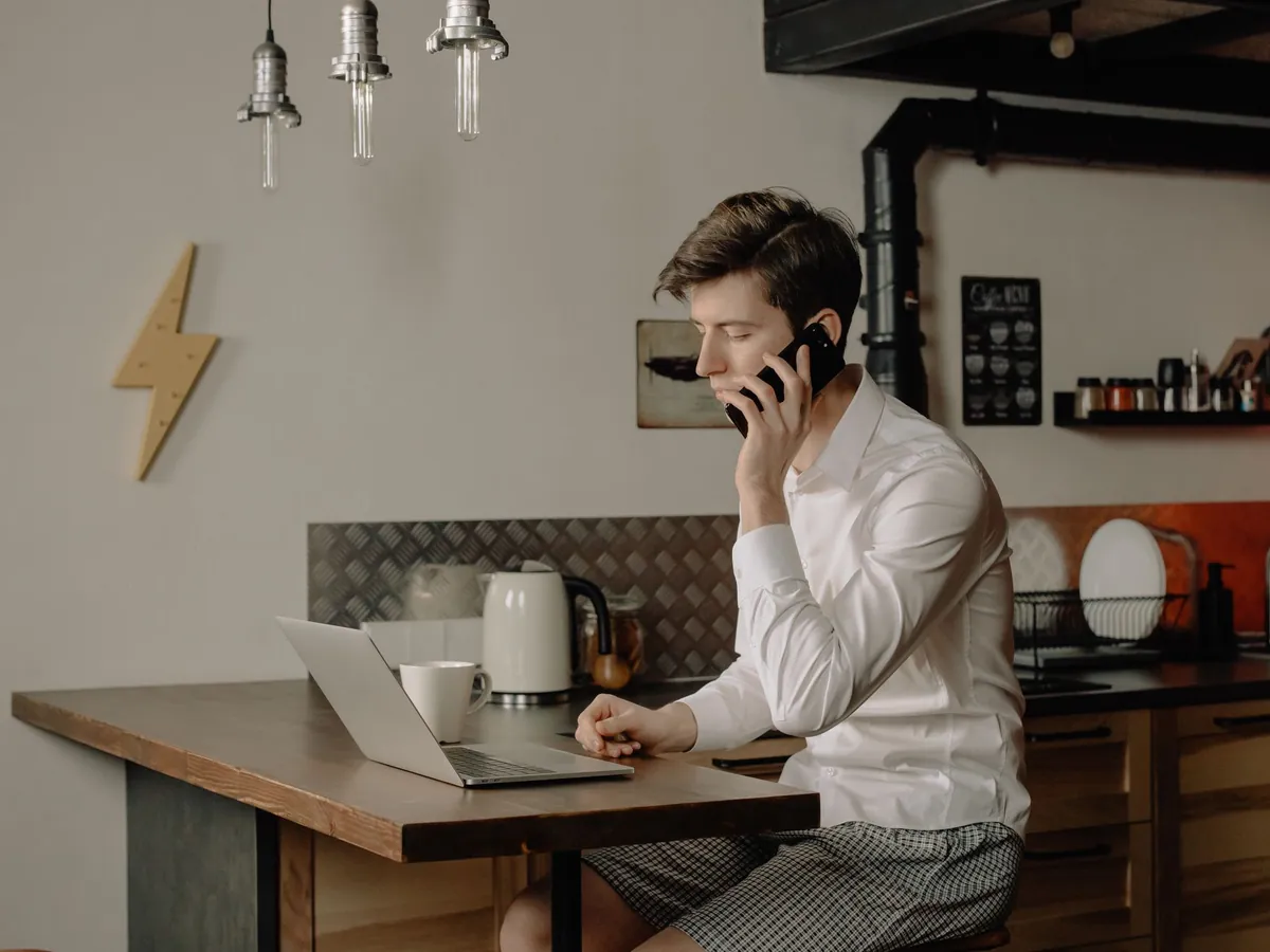 Young man working at a home office desk, talking on a phone while using a laptop