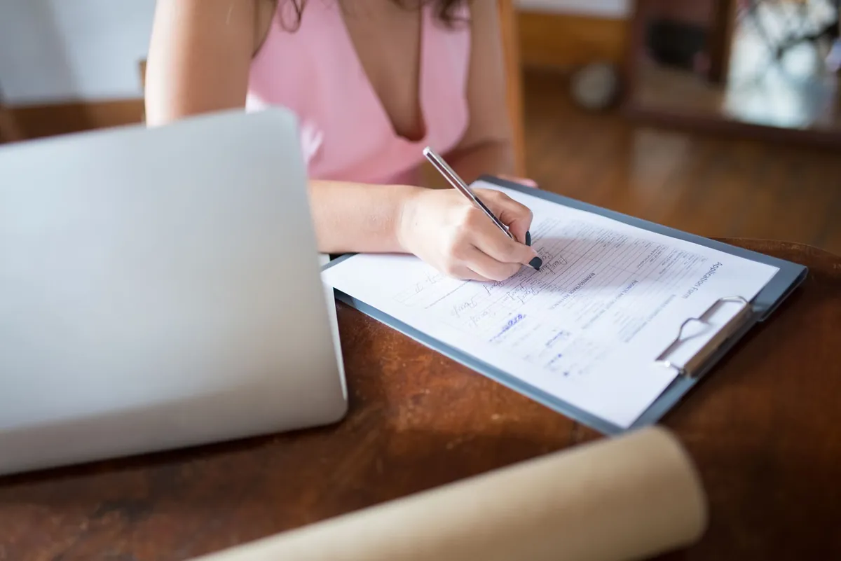 Woman writing on a document on a clipboard at a wooden desk with a laptop nearby.