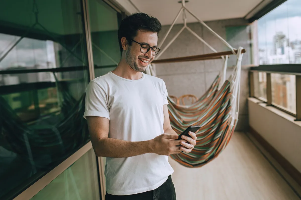 Smiling man using his smartphone on a balcony with a hammock.
