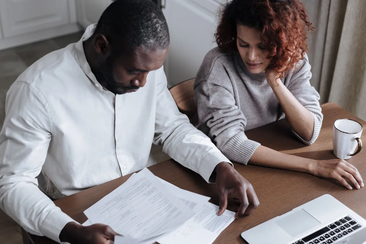 Two people sitting at a home desk reviewing documents beside a laptop