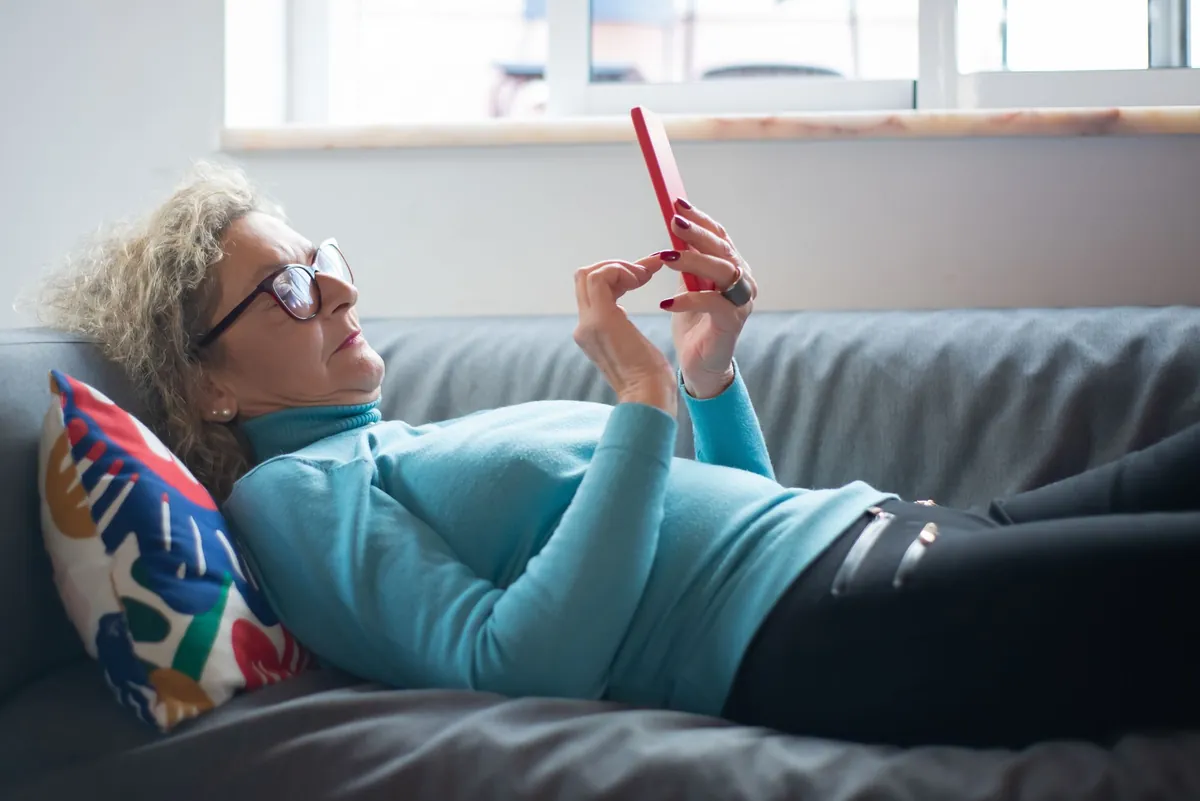 Older woman with glasses lying on a grey sofa looking at a red phone