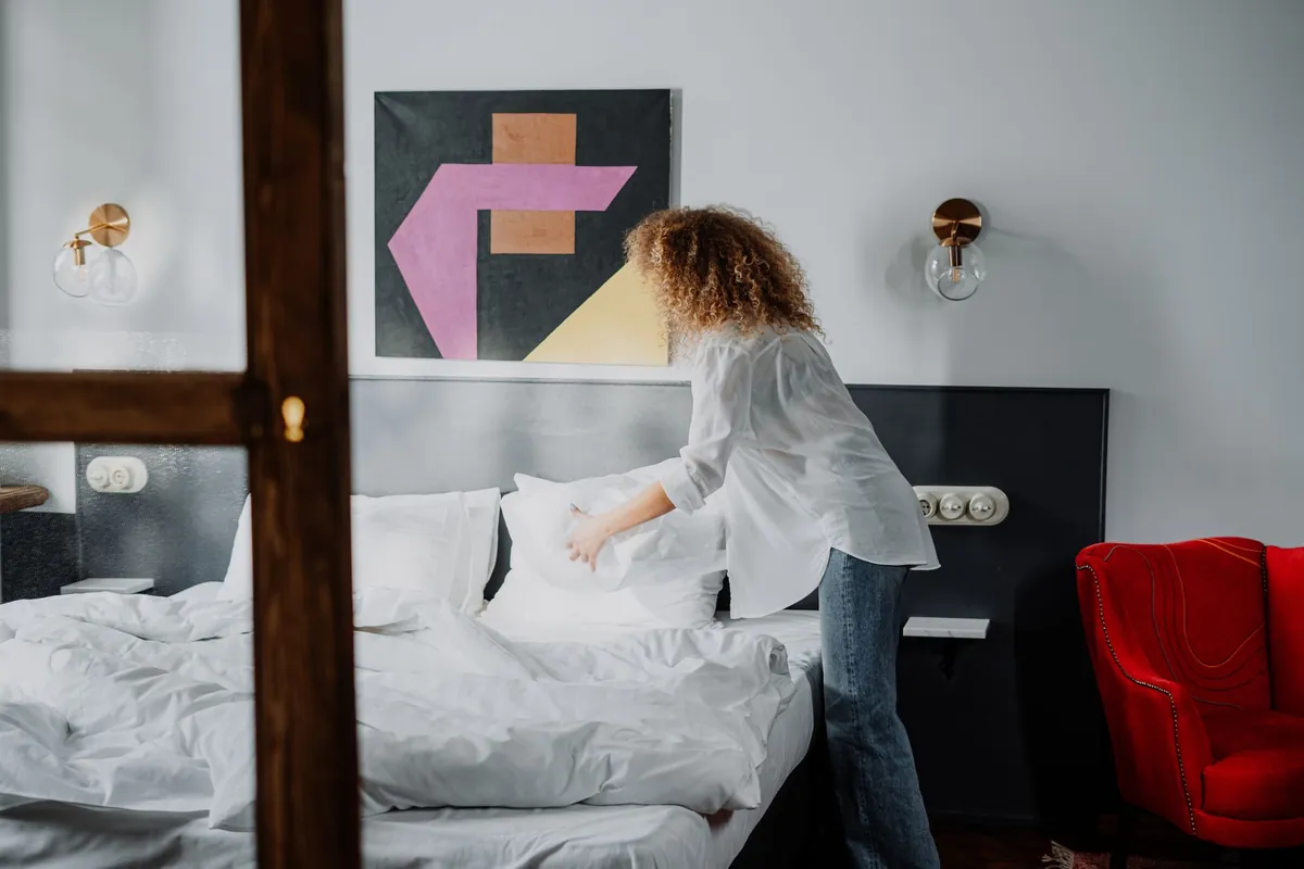 A woman with curly hair making a bed with white linen in a bright bedroom