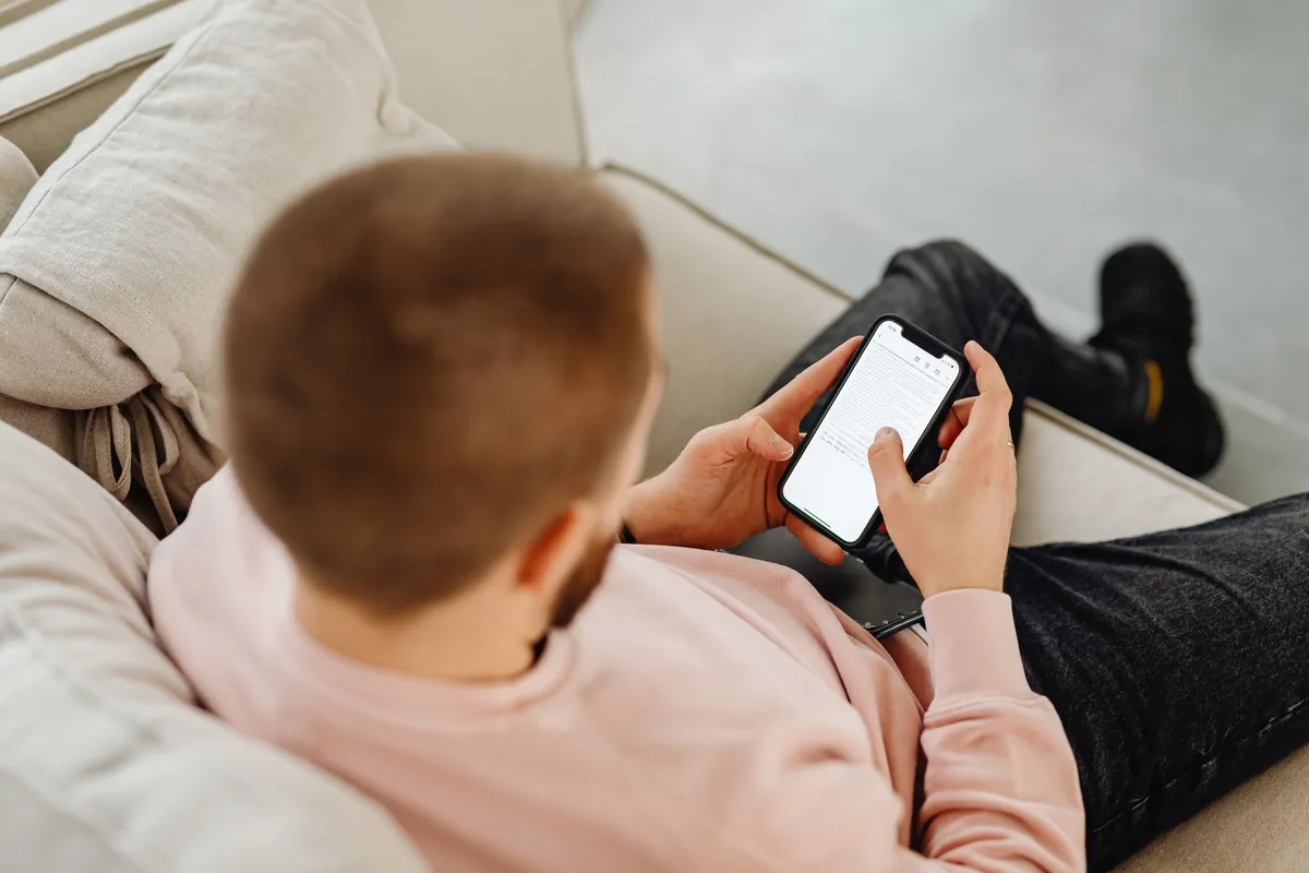 A man sitting on a sofa indoors, looking at his smartphone in a bright living room.