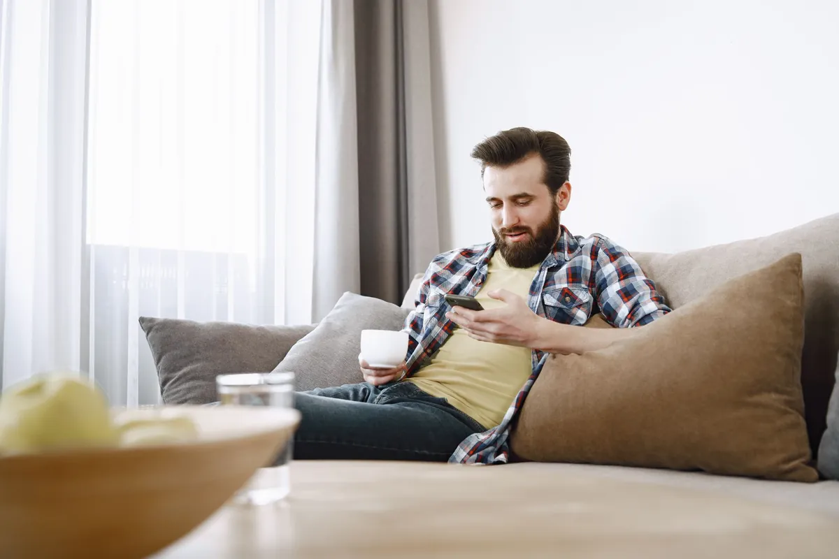Man sitting on a sofa using a smartphone with a cup of coffee nearby