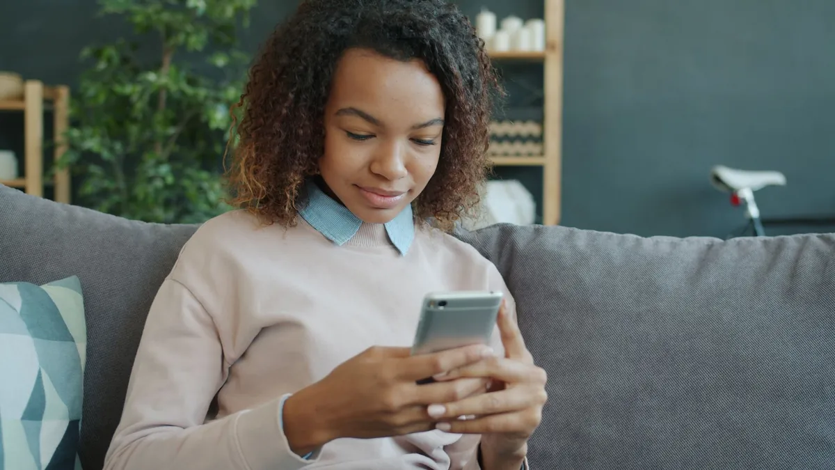 A young woman sits on a couch indoors, looking at her smartphone.