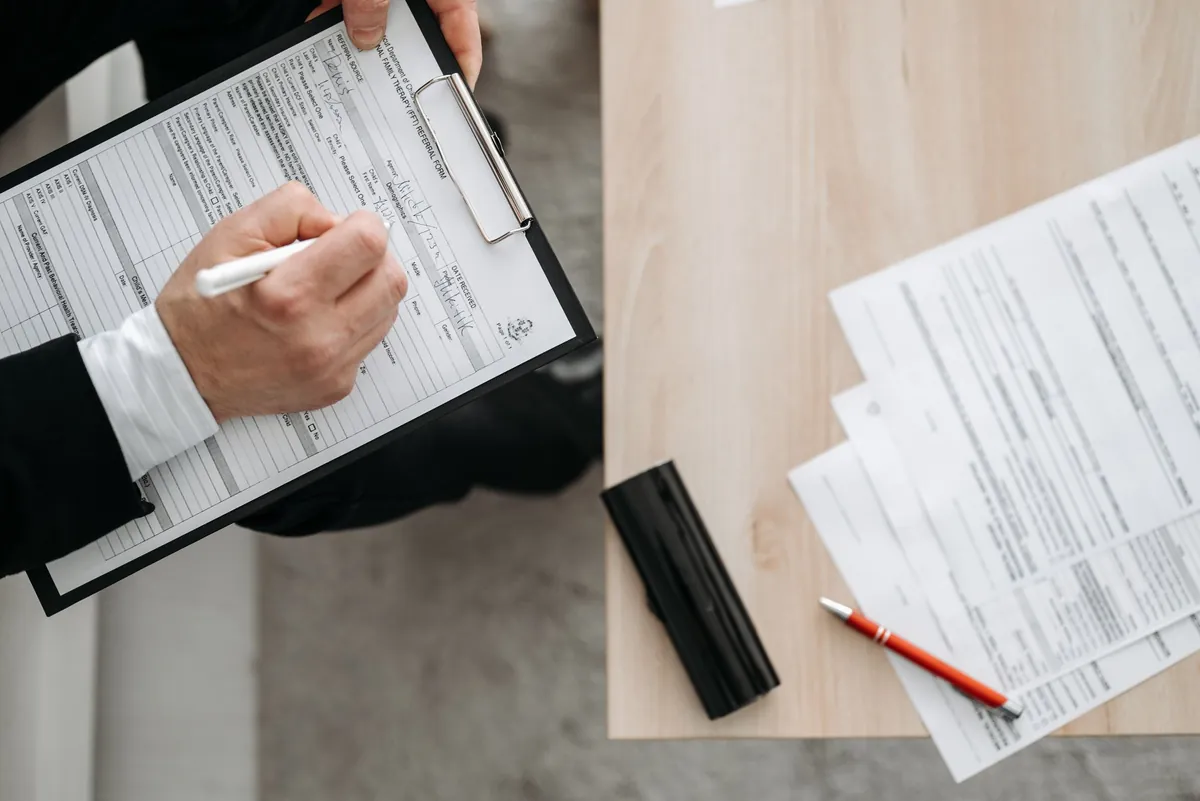 Person filling out paper forms on a clipboard viewed from above at a desk