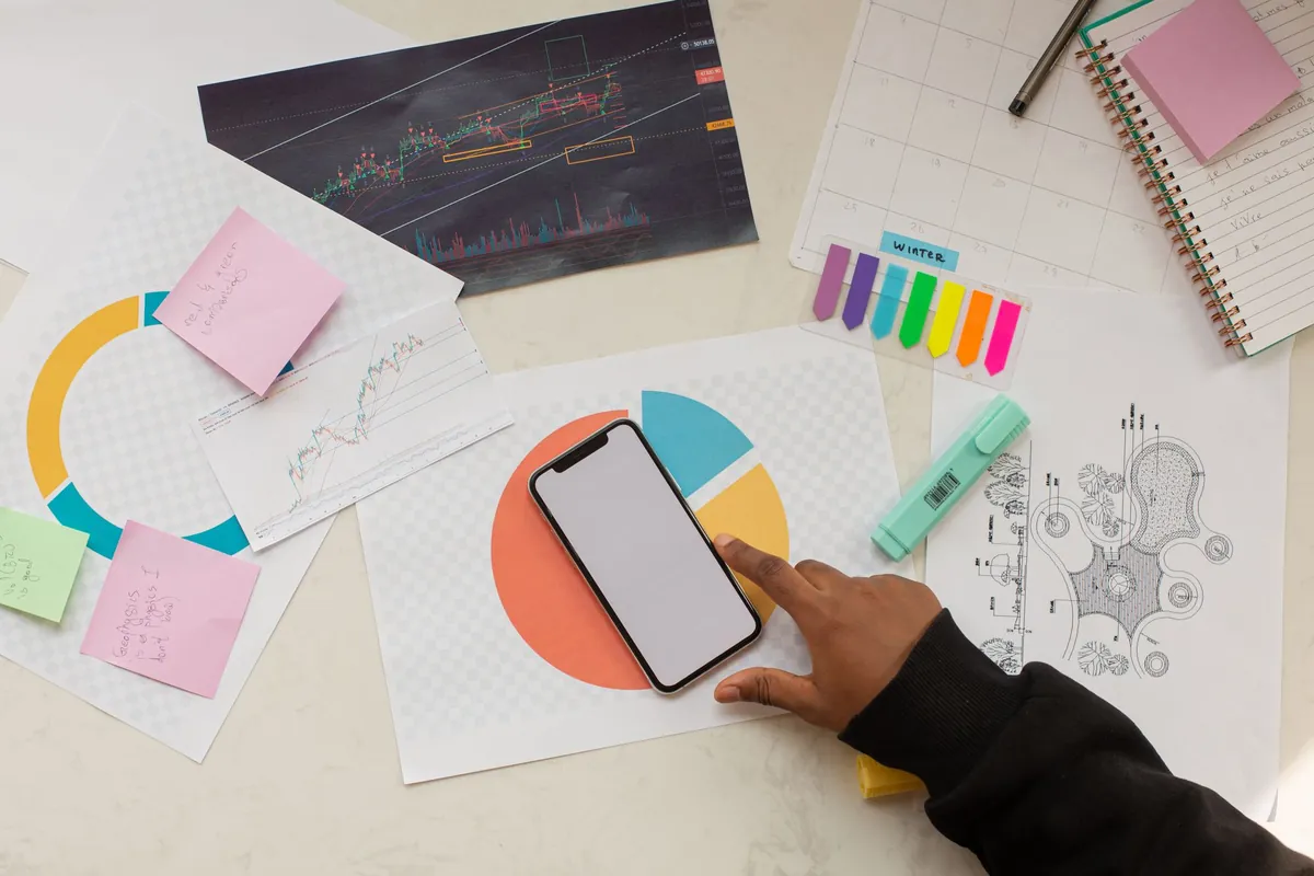 A hand touches a smartphone on a desk with printed charts, notes, and a notebook.
