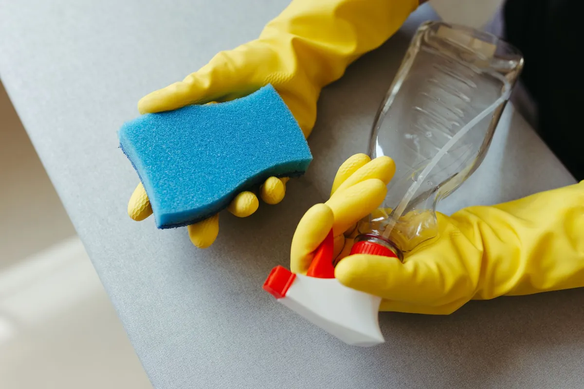 Yellow-gloved hands hold a blue sponge and a spray bottle above a countertop.