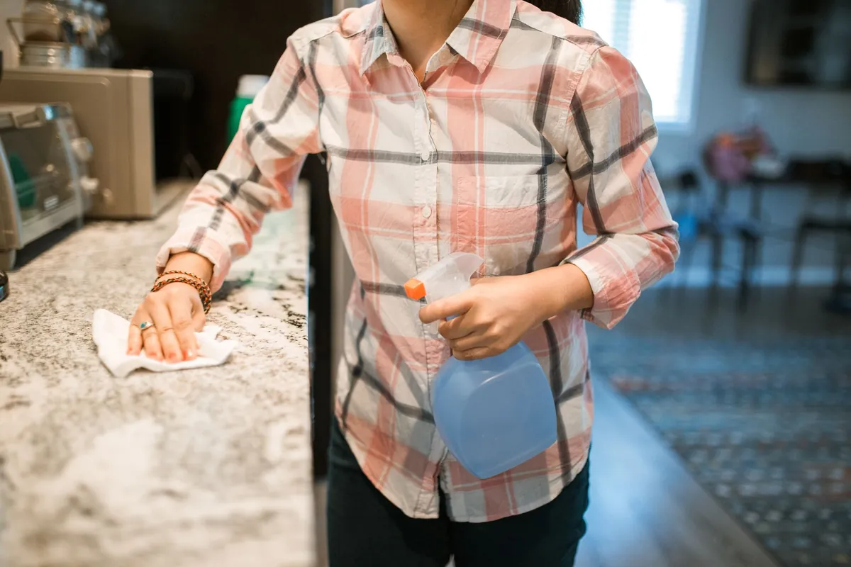 Woman in a plaid shirt wiping a kitchen counter with a spray bottle and cloth