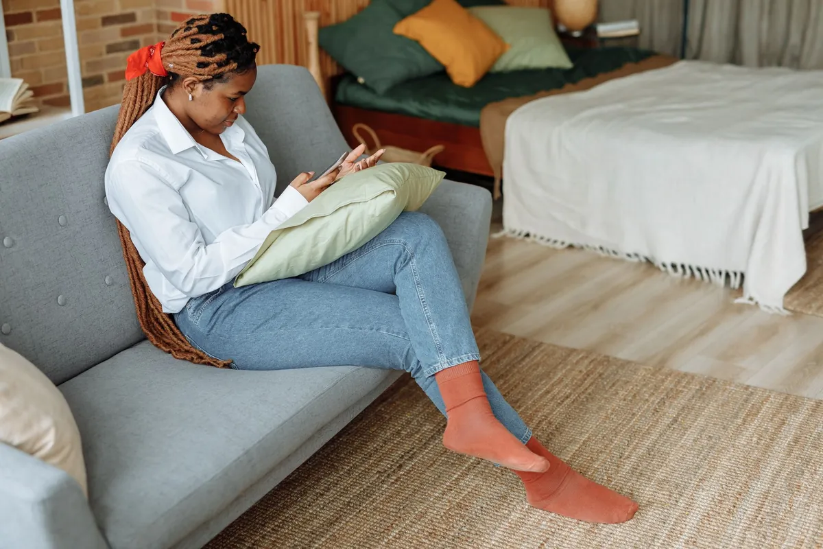 Woman with dreadlocks sitting on a sofa at home, looking at her cellphone