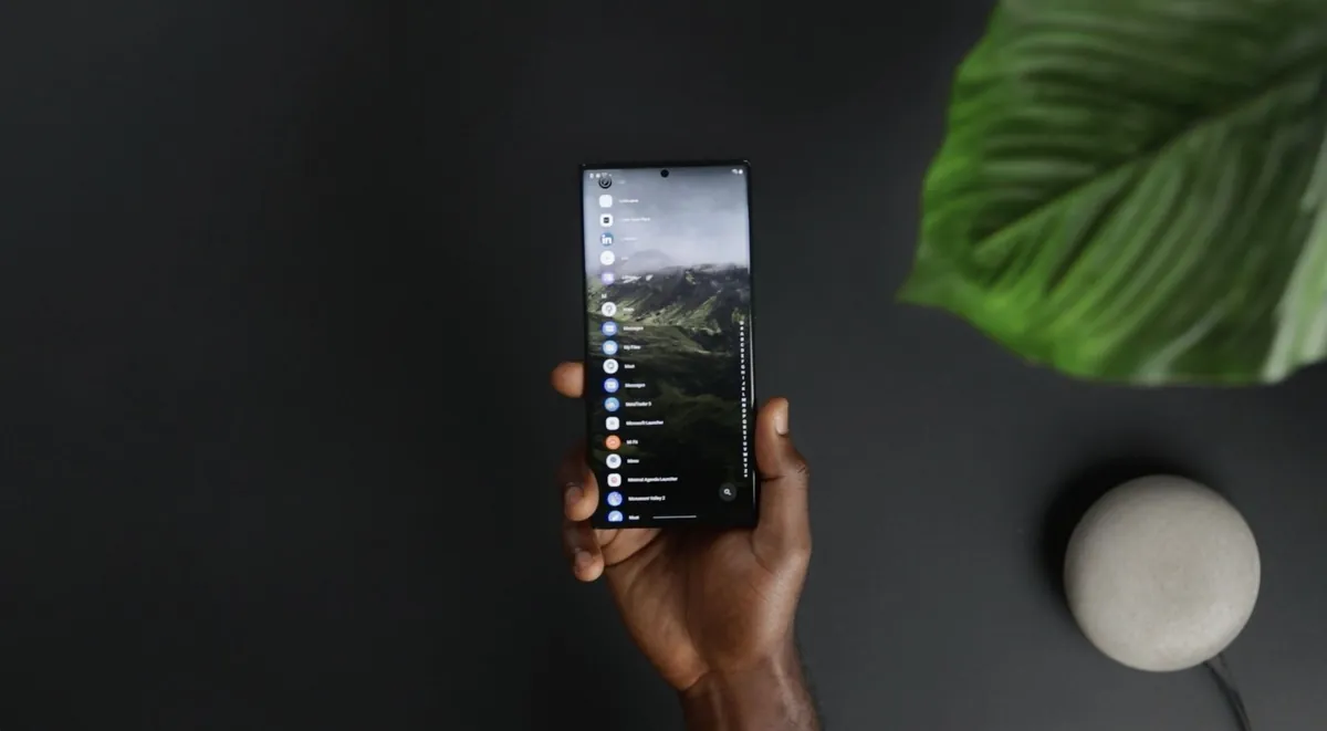 Hand holding a glowing smartphone in a dark room near a green leaf and a small round speaker