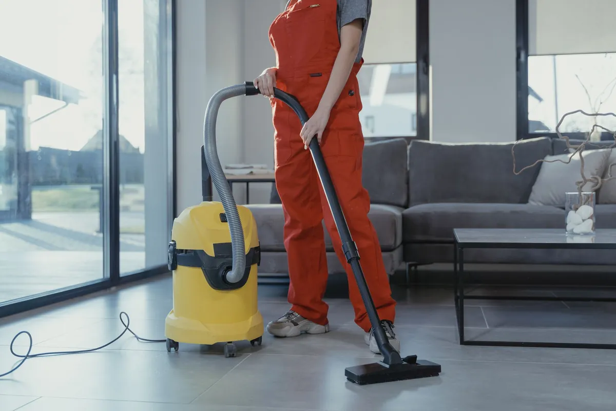 Cleaner in an orange uniform vacuuming the floor of a bright living room with a grey sofa