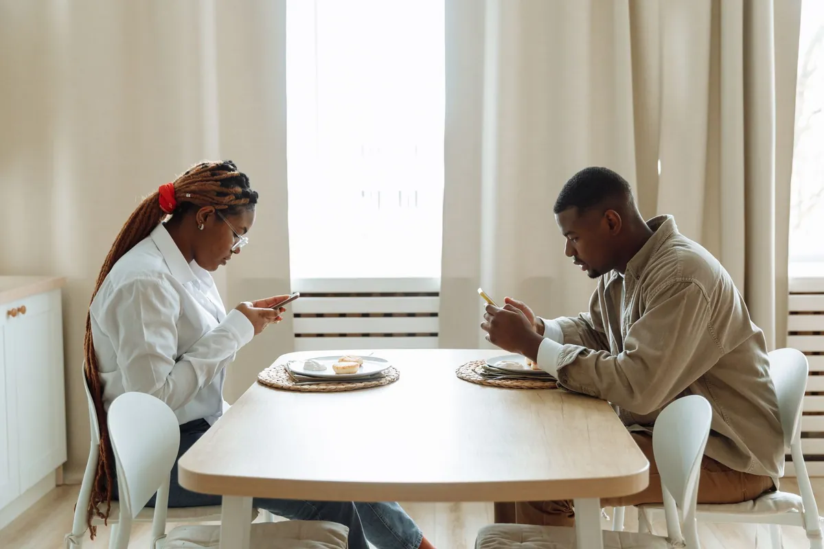 Two adults sitting at a dining table indoors, each using a smartphone