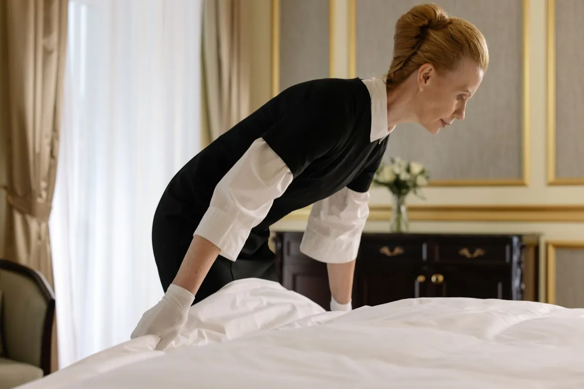 Housekeeper in uniform and gloves making a bed in an elegant hotel-style room
