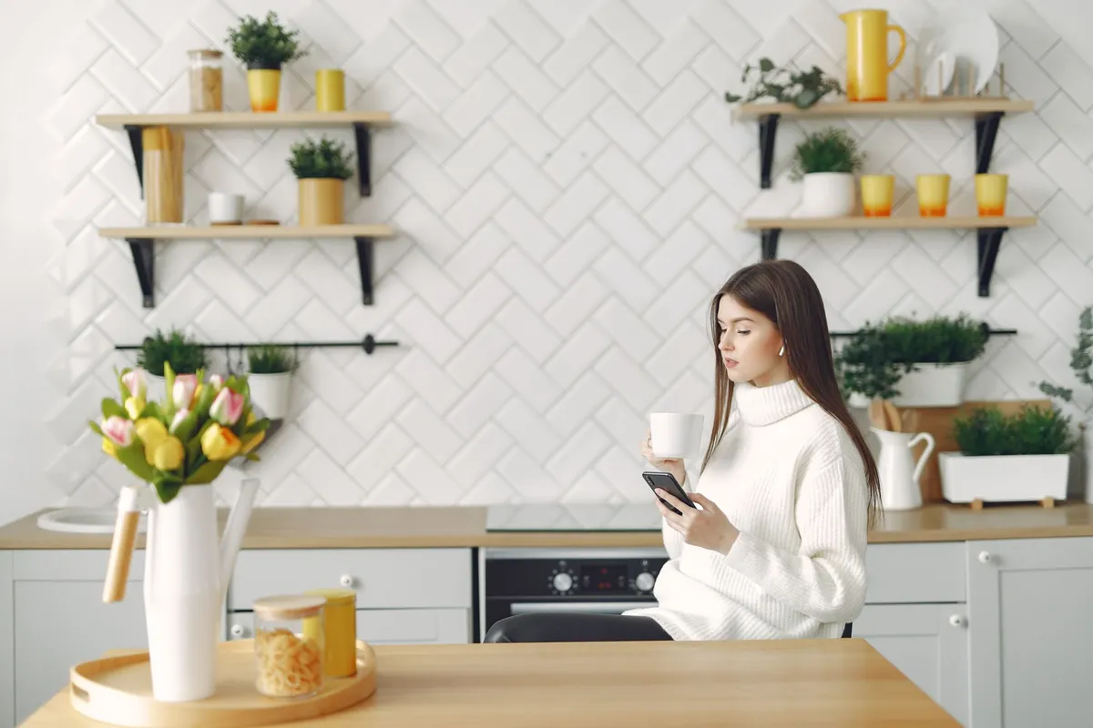 Woman in a warm sweater browsing her phone at a kitchen table with tulips and a hot drink