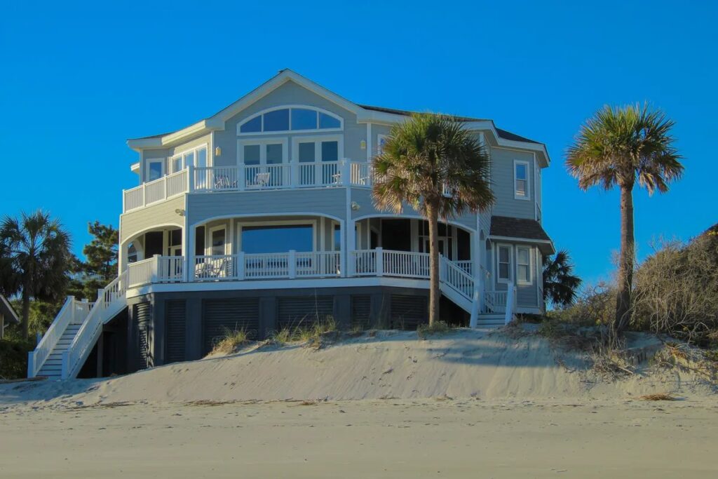 Pale-yellow two-storey beach house on stilts with white railings and palm trees on a sand dune