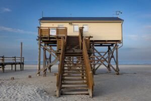 Beach house on tall wooden stilts with a long staircase leading down to sand under a blue sky