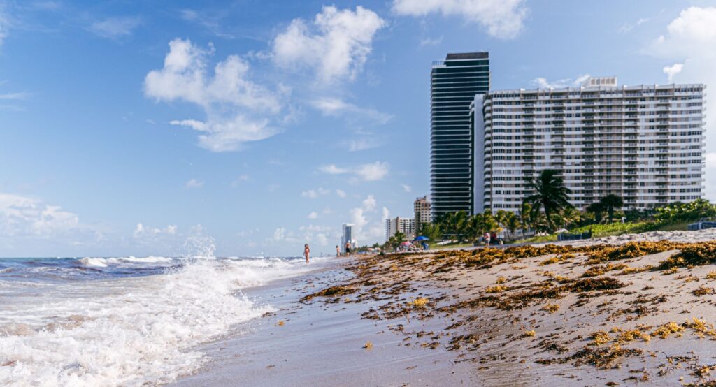 Waves rolling onto a sandy beach beside modern buildings under a blue sky