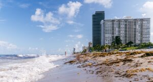 Waves rolling onto a sandy beach beside modern buildings under a blue sky