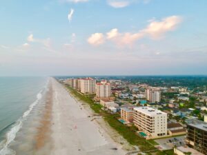 Aerial view of a beach town with a wide sandy shoreline, ocean, and low-rise buildings under a bright sky.