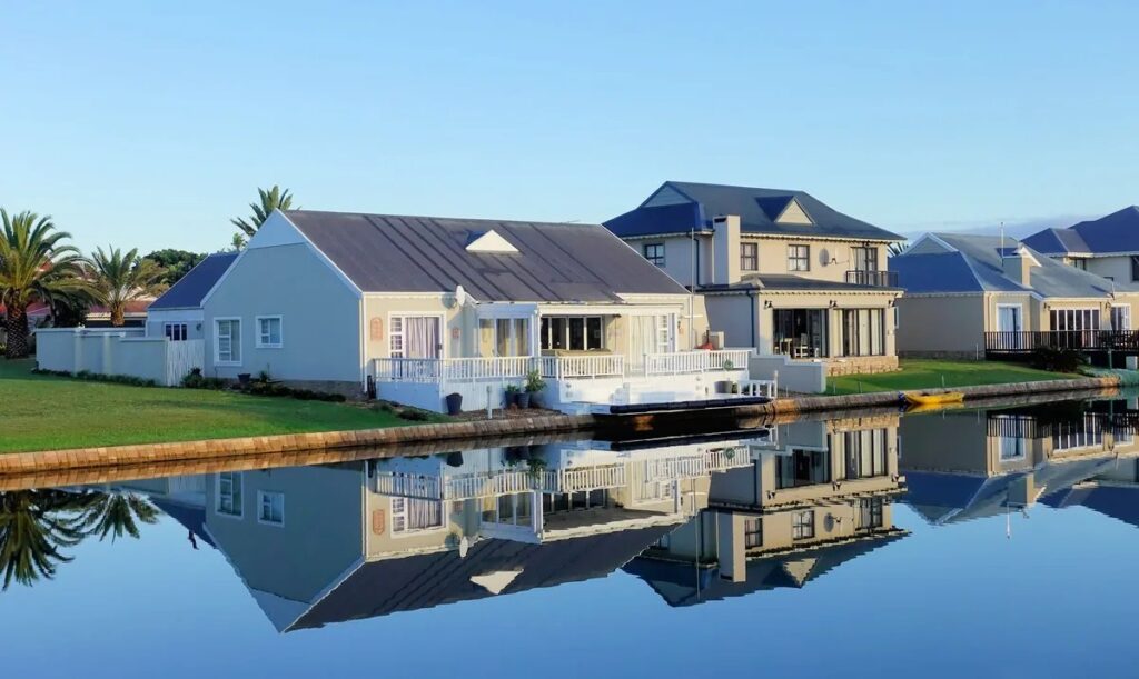Row of waterfront homes with grey roofs and palm trees reflected in a calm canal under a blue sky
