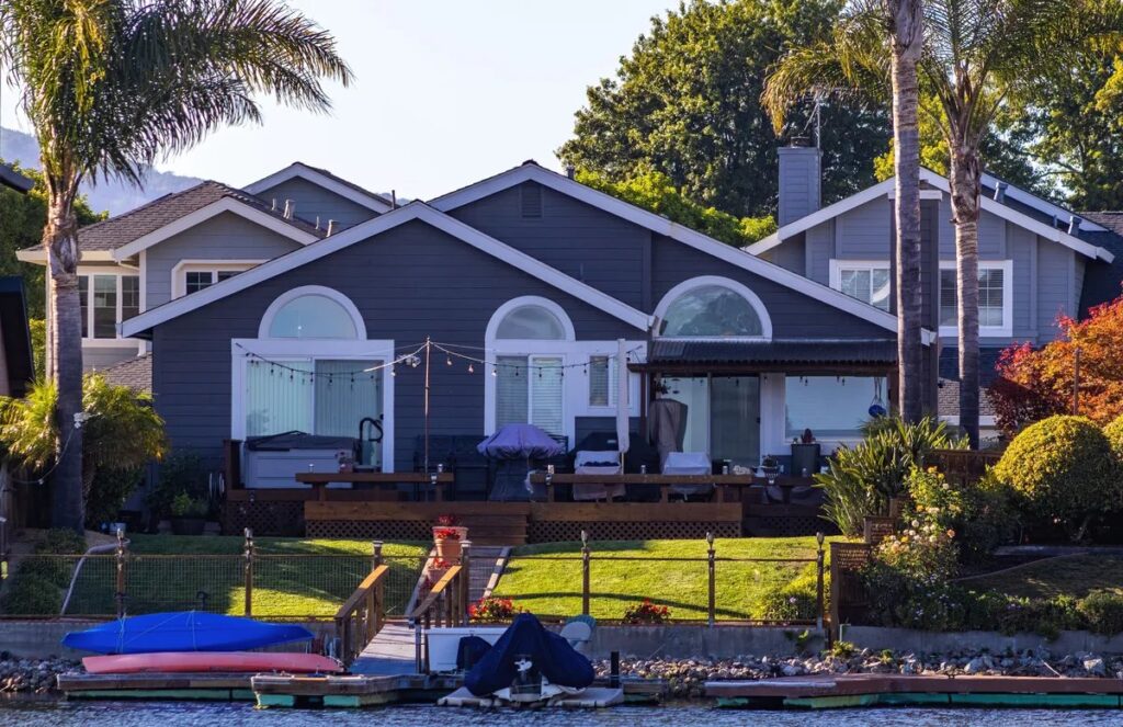 Grey waterfront house with palm trees, lawn, and kayaks on a dock