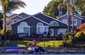 Grey waterfront house with palm trees, lawn, and kayaks on a dock