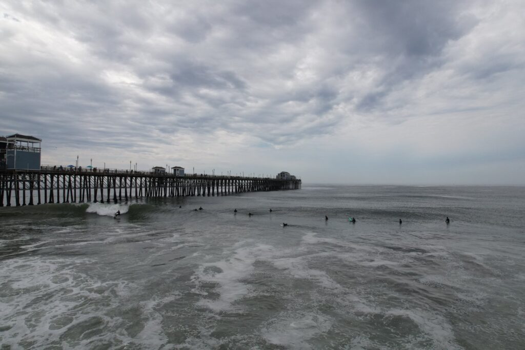 Surfers in the water beside a long wooden pier under a cloudy sky.