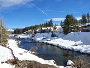 Snow-covered houses beside a river with pine trees under a blue sky.