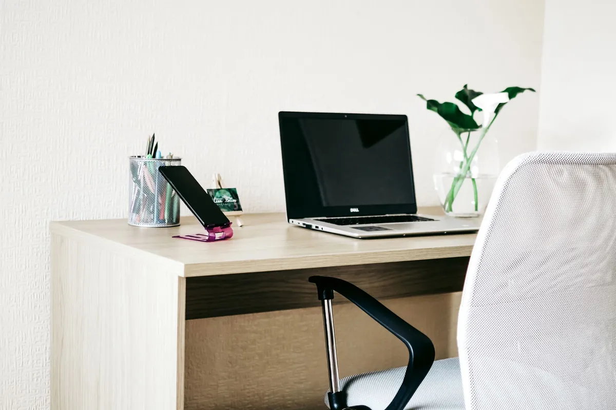 White desk with laptop, phone on a stand, and small plant by a window