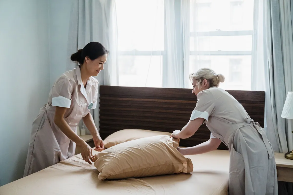 Two housekeepers in uniforms making a bed and fluffing pillows in a bright bedroom